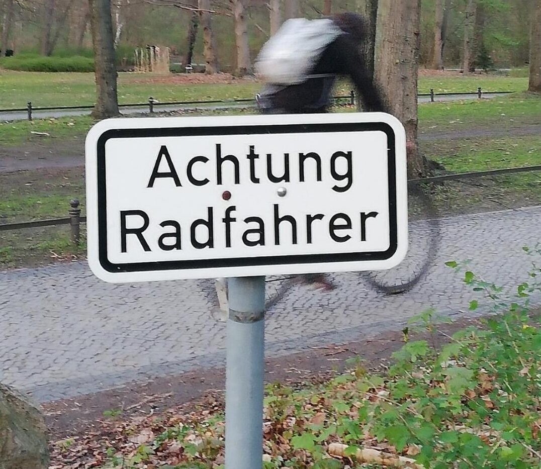 Radfahrer fährt schnell durch einen Park. Davor ein Schild: Achtung Radfahrer!