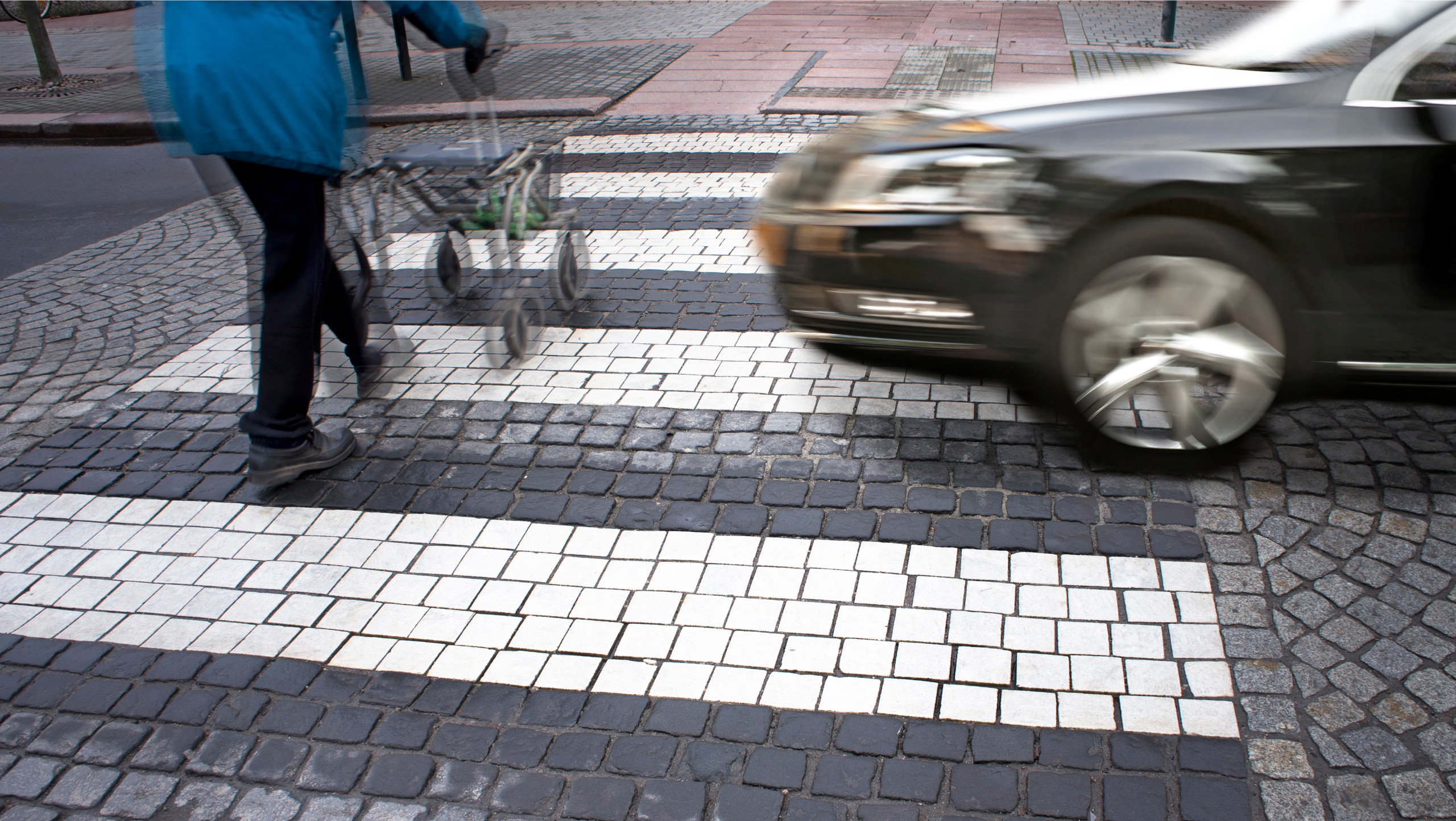 ältere Person mit Rollator überquert die Straße auf einem Zebrastreifen während ein schnelles Auto sich nähert