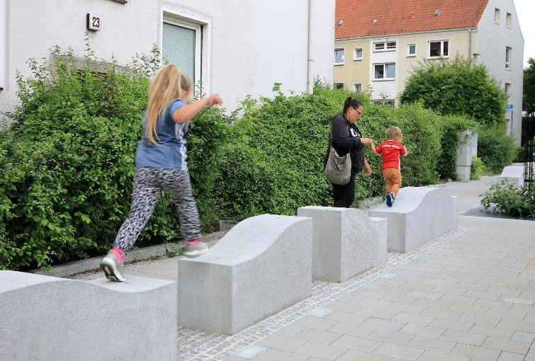 Kinder laufen über wellenförmige Spielelemente aus Beton entlang eines Gehweges.